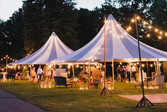 Outdoor event with large white tents and string lights and speakers in a park setting.