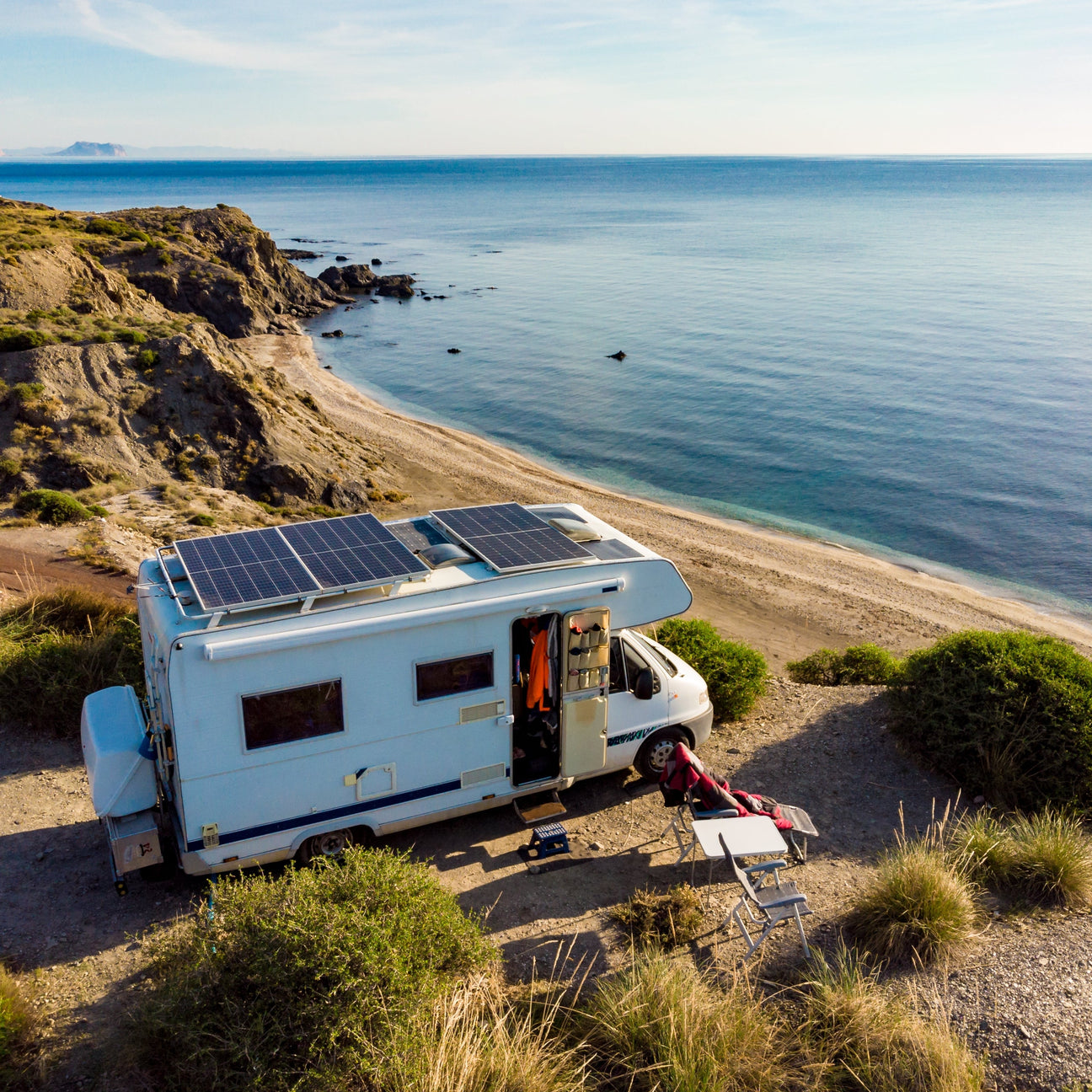 An RV with rigid solar panels installed on top, parked by the ocean.