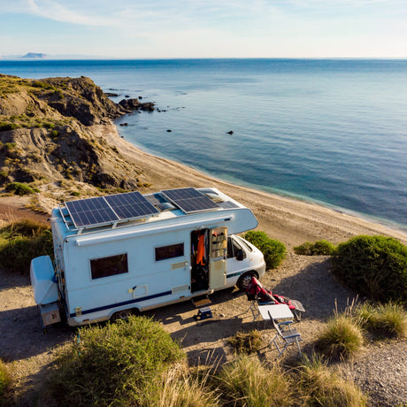 An RV with rigid solar panels installed on top, parked by the ocean.